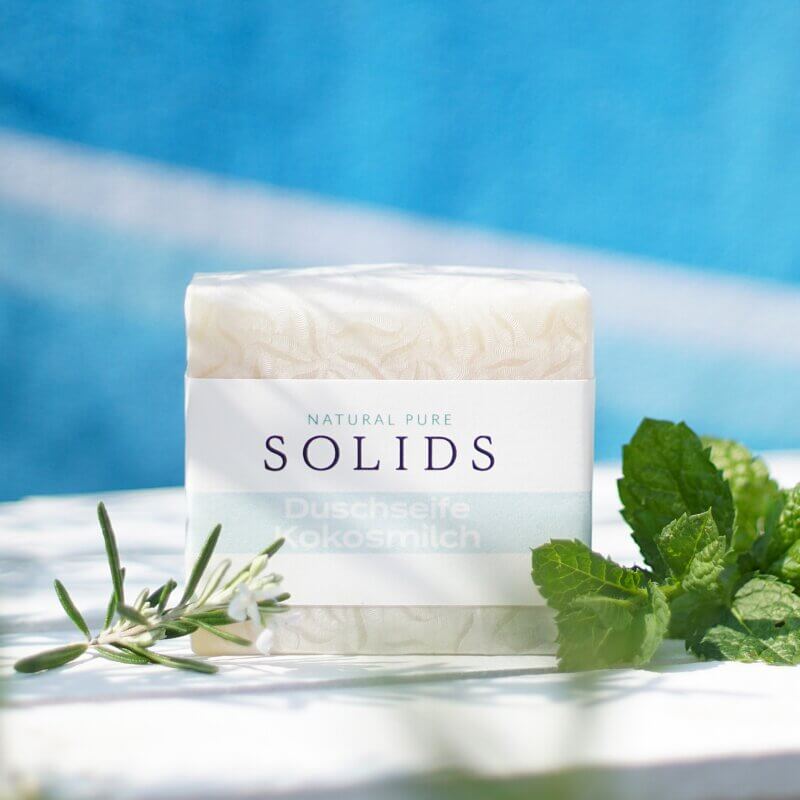 Duschseife Kokosmilch mit Rosmarin und Minze auf weißem Holz und blauem Hintergrund von Natural Pure Solids, Vegane Milchseife mit Verpackung und natürlichen Kräutern